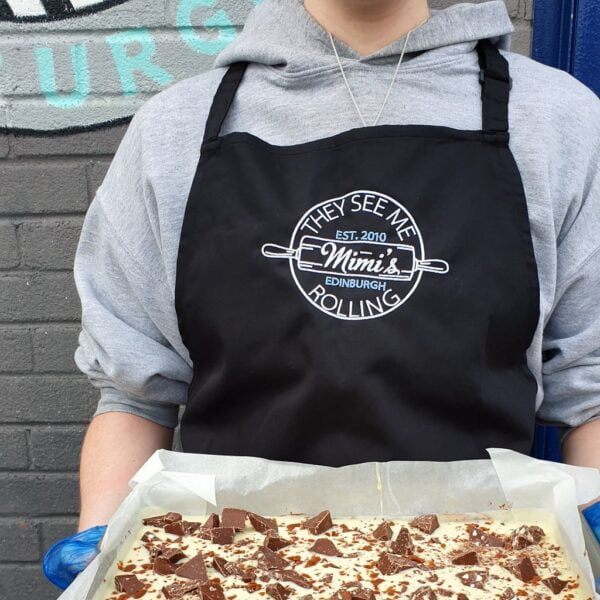 A person in a Mimi's Bakehouse apron holding a traybake cake